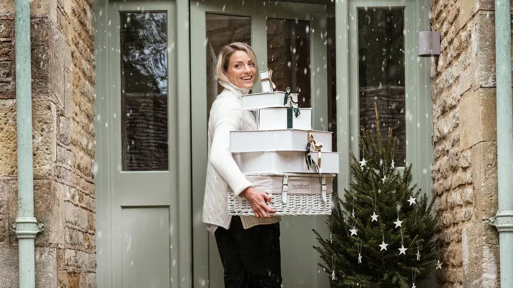 A woman stands on the steps of a front door holding a pile of presents