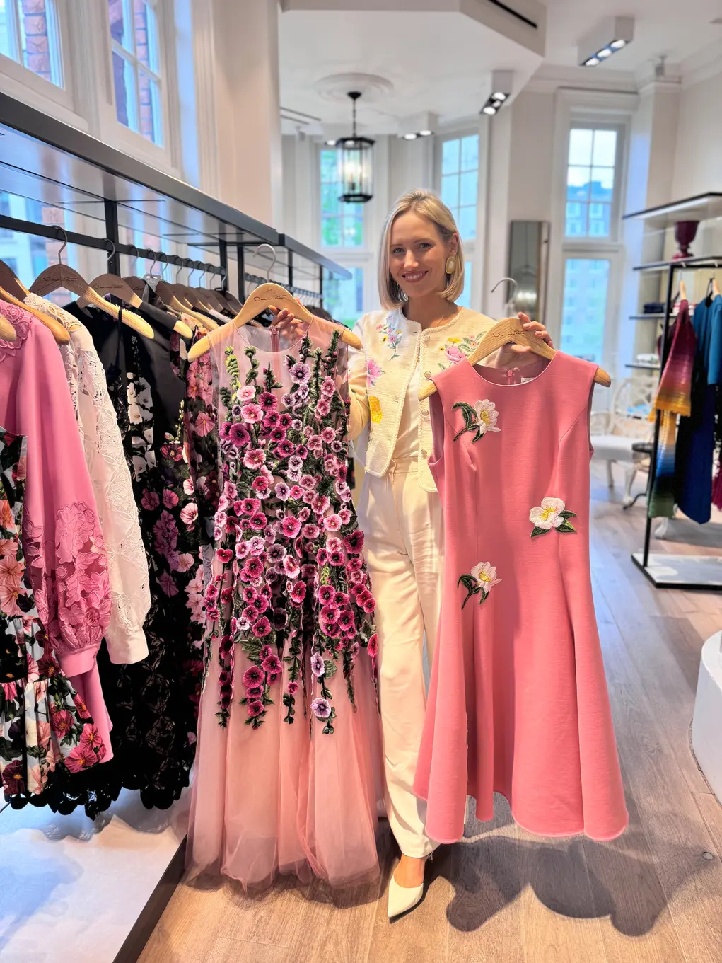 Blonde woman in white dress holds up a pink dress and a pink floral dress