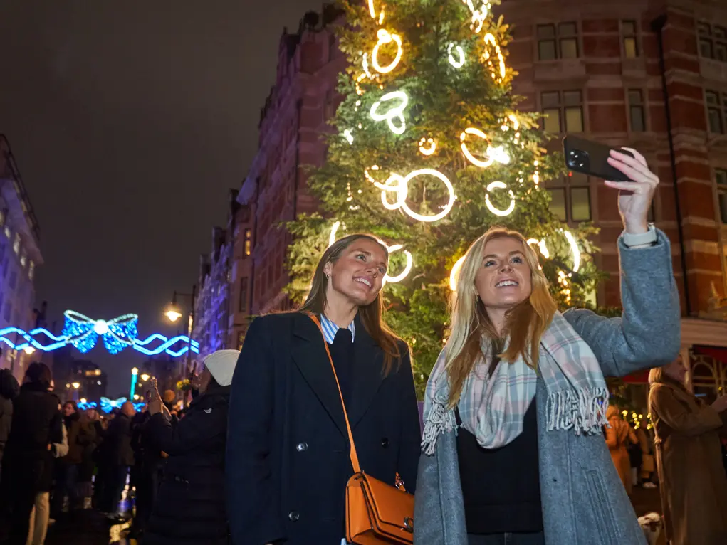 Two women take a selfie in front of a large Christmas tree